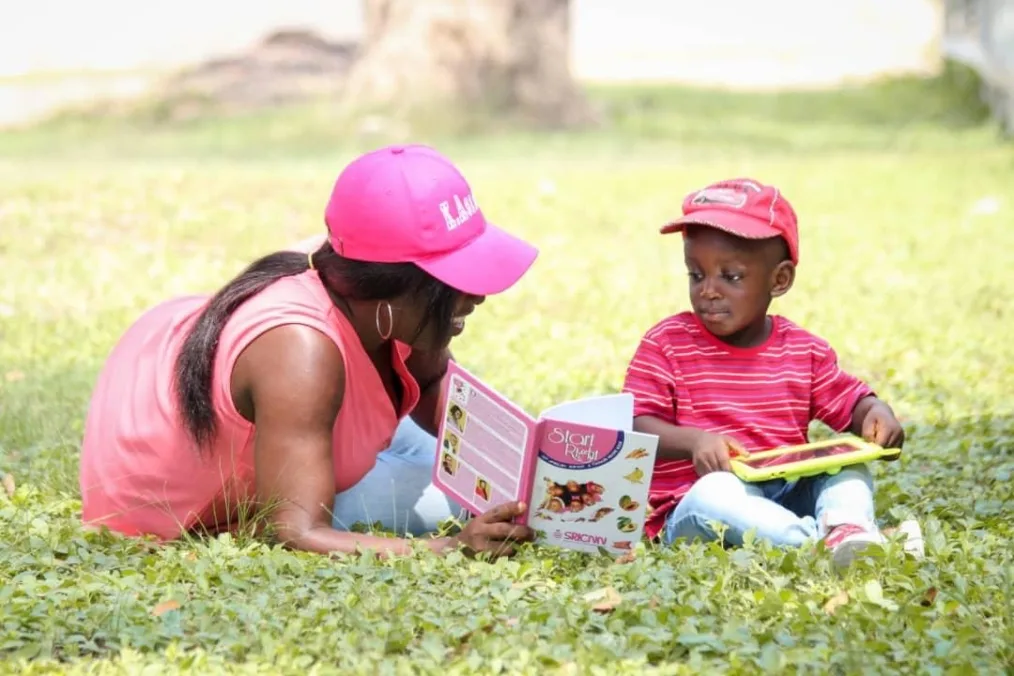 Obama Leader Vera Osei-Bonsu reads her children’s recipe book to a small child while laying in the grass on a sunny day. Both are wearing baseball caps..