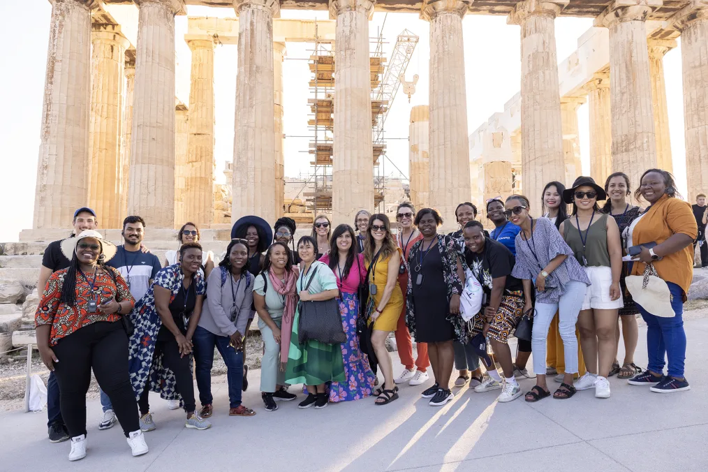 A group of 24 people stand together in a group photo in front of the Acropolis in Athens, Greece. It is sunny outside. They are all of different ages and ethnicities. They have a range of light to deep skin tones. Columns and construction are in the background. 