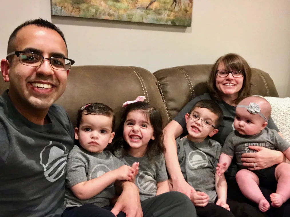 A man and woman sit on a brown couch with four young children between them. All wear gray t-shirts with the Obama sunrise logo.