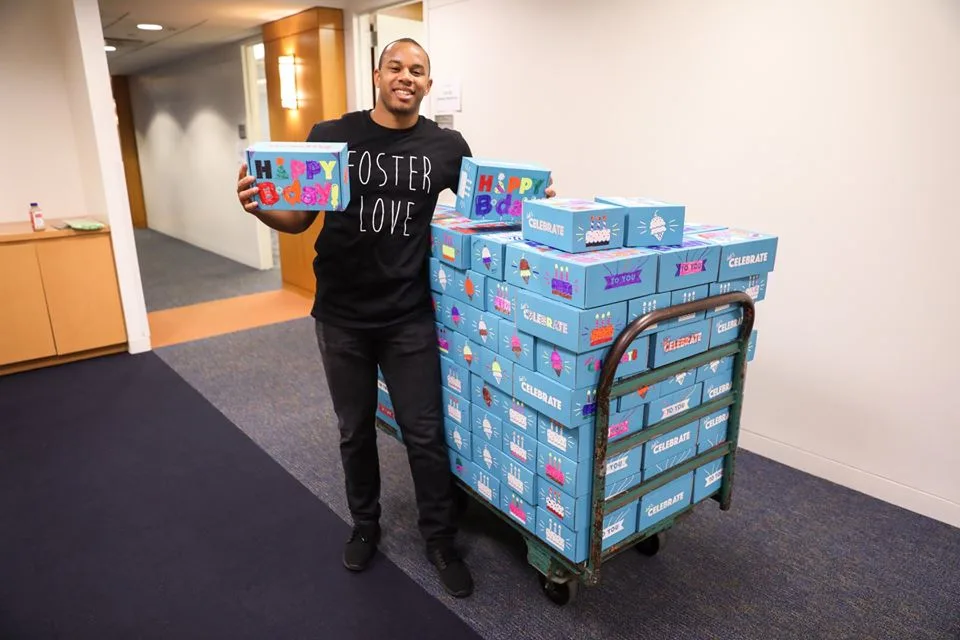 A man smiles next to a large cart holding dozens of birthday boxes for children in need.