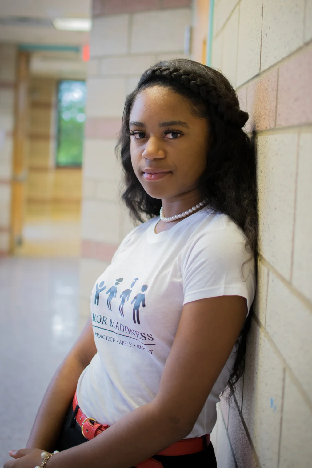A medium skin toned Black woman is leaning to the right against a wall smiling with her mouth closed. She has long dark hair with a braid in the front and is wearing a white T-shirt, red belt and a pearl necklace. Her arms are crossed in front of her; the background appears to be a hallway. 