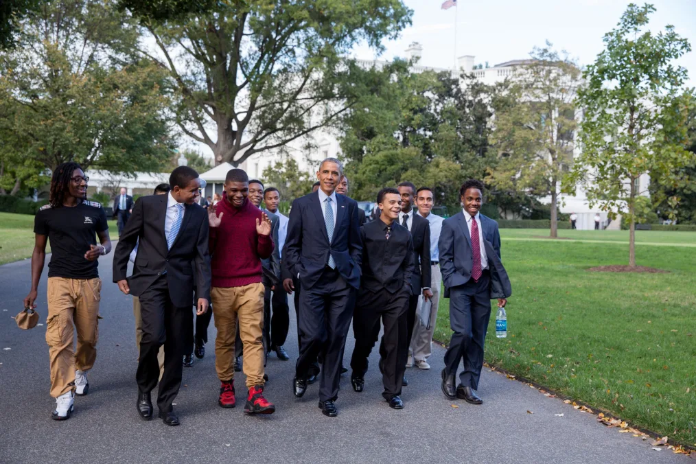 On the South Lawn, President Obama walks in the middle of a group of young men. All have a range of light to deep skin tones. They are wearing a mix of casual and professional attire. 