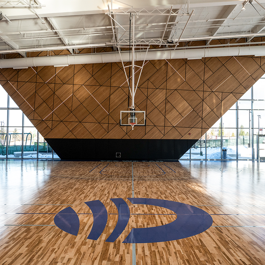 An image of Main Court, the basketball court located in Home Court. The image features the wood net-themed basketball backboard, the basketball net, and the basketball court floor. 