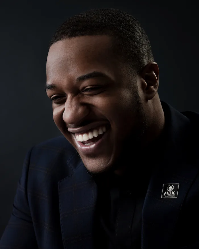 A portrait photo of a man with a deep skin tone wearing a navy blue and tan plaid blazer with white 'MBK Alliance" stitching on the chest smiles toward the left-hand side of the photo in front of a gradient black background. 