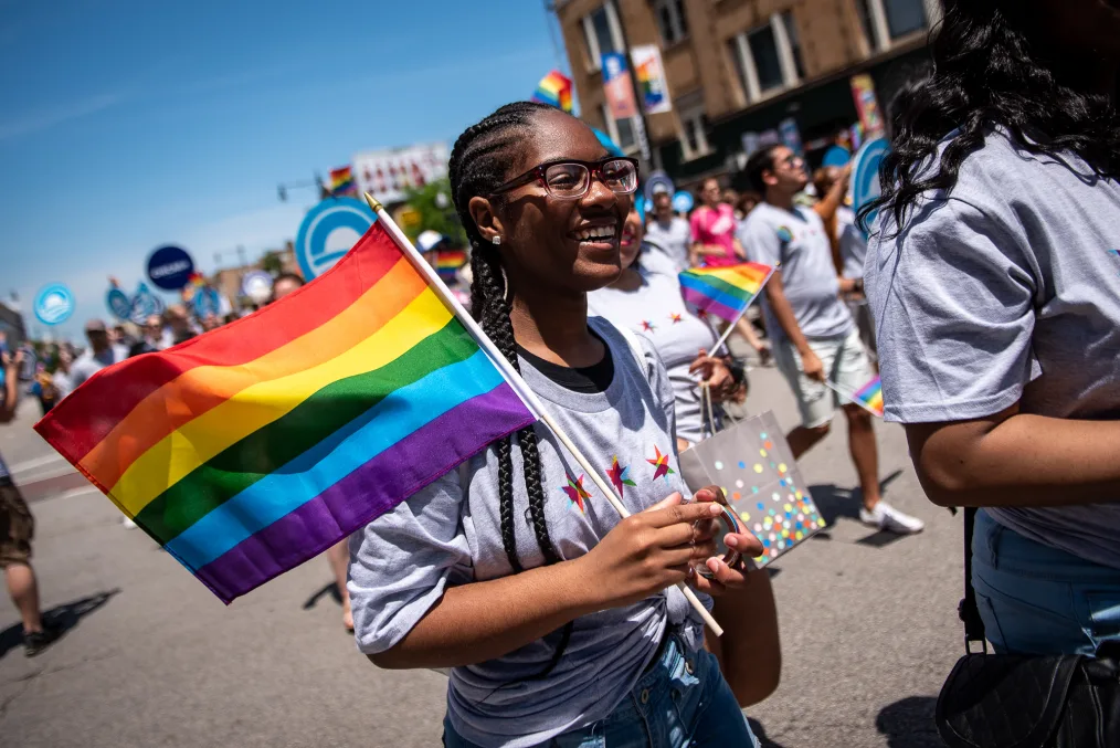 
The camera focuses on a young girl with a deep skin tone, braids, and glasses holding a pride-colored flag while walking in the pride parade  