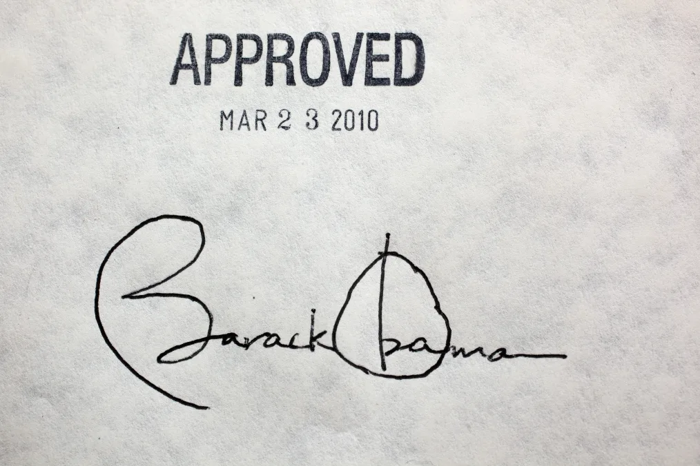 The image is a photograph of Barack Obama’s signature signed on the Affordable Care Act. The image is of a sheet of paper: at the top of the paper is the word “Approved” stamped on. Underneath “Approved” is text that reads, “March 23 2010.” Below that is President Barack Obama’s signature. 