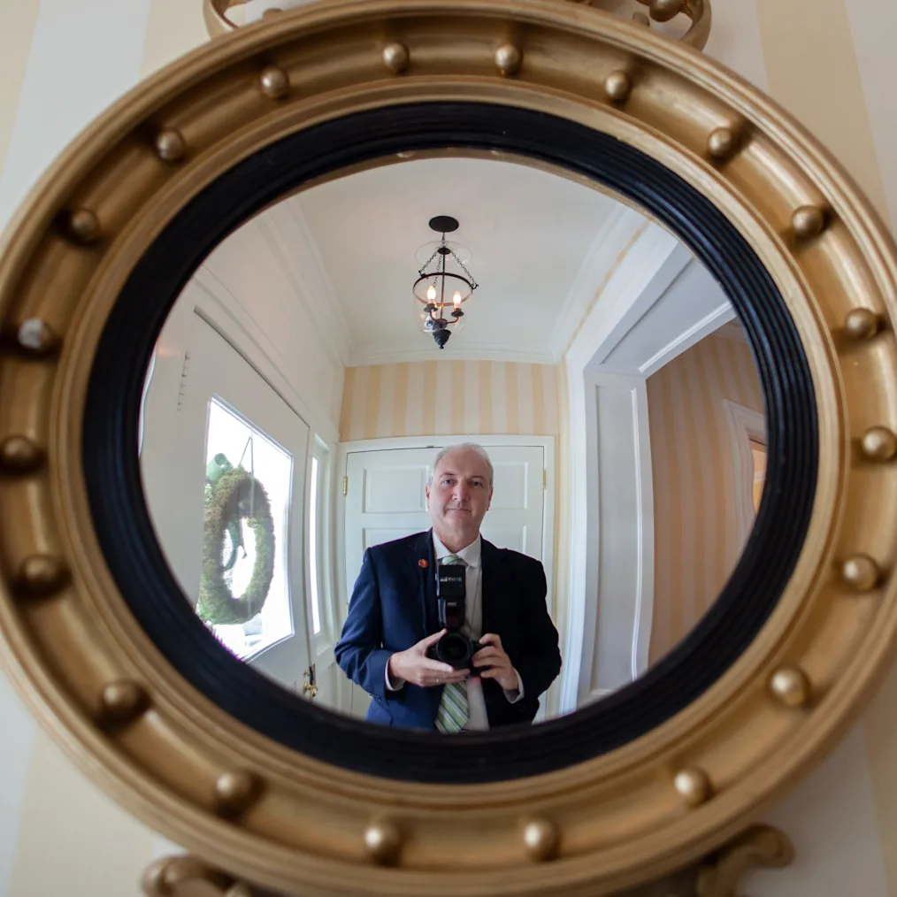 Chuck Kennedy, a man with a light skin tone smiles as he stands in a gold circle mirror. 
