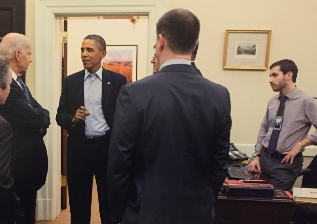 The image is located in a room in the White House. In the background of the image is an open door, a dark brown desk with a computer and files on top of it, and a beige wall with a square picture and a gold frame. In the center of the image are six men standing in a circle and one man standing behind the desk. In the six man circle, President Obama stands in the center. He is wearing a dark colored suit and a light blue button-down shirt. On the lapel of his suit is an American flag pin. He is in mid conversation. Surrounding him are five men, four of the men are indistinguishable because their faces are turned away from the camera. One of the men whose profile is towards the camera is identified as current President Joe Biden. He is wearing a blue polka dotted tie and standing with his arms crossed against his chest. The man – outside of the circle, standing behind the desk – is Craig Dorsett. Craig is standing profile to the camera staring at the conversation between the circle of men. He has one hand on his hip and another on the desk. He is a pale man with short dark brown hair and a dark brown goatee. He is wearing a light gray button-down shirt, a dark color tie, dark pants, and a watch on his right arm. On his neck is a White House ID, hanging on a chain. 