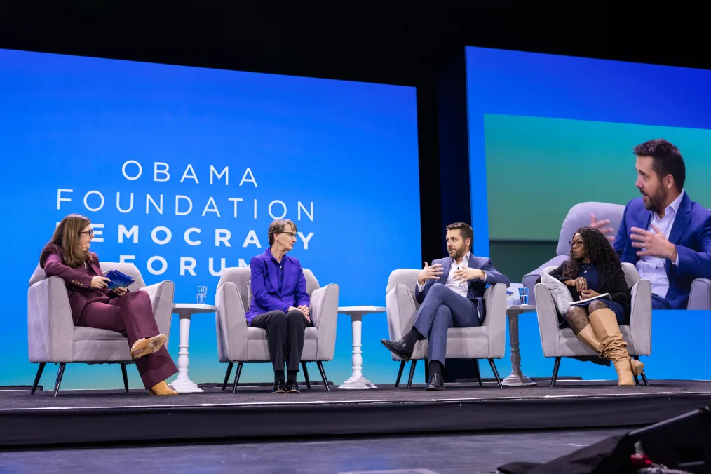 Brian Deese speaks on a panel alongside Mary Kay Henry, Chiedo Nwankwor, and Betsey Stevenson. All speakers are a range of light to deep skin tones and they sit on a stage. A screen behind them reads, “Obama Foundation Democracy Forum.