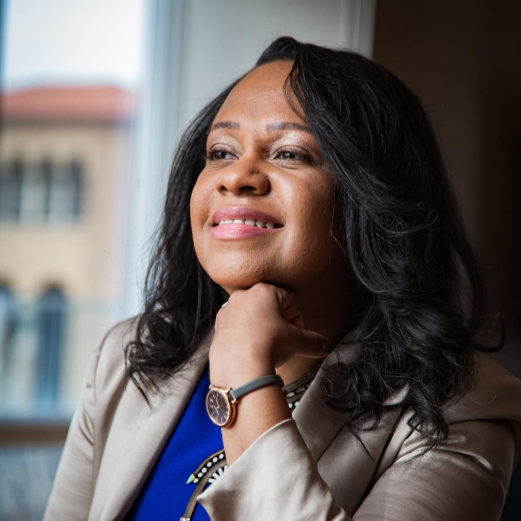 The image is a headshot of Ny Whitaker. Ny has a medium deep complexion, long black hair, and dark brown eyes. She is wearing a gold blazer, a blue shirt, and a black watch with a gold frame. She is smiling off into the distance looking away from the camera to the left side. Behind her is a brown wall and a window that overlooks a city building.