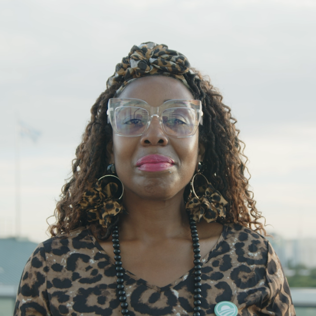 The image is a gif of Patrician Haywood smiling. Patrician has deep skin complexion and long dark curly hair. She is wearing square lucite glasses, a leopard headwrap, leopard hoop earrings, a leopard blouse, and a black beaded necklace. On the right side of her shirt is an Obama Foundation "Rising Sun" logo pin in the color blue. She is standing outside in the daytime with the Chicago skyline in the background.