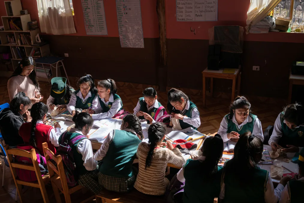 A group of 13 indigenous girls work together at a long table with natural light streaming through the educational center’s windows and their tutor looking on. The girls are wearing school uniforms with a collared shirt, green sweater vest, and plaid skirts.