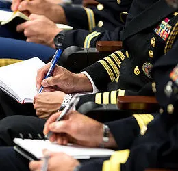 A few pairs of hands with a light-medium skin tone are writing in notepads. They are dressed in uniform wearing black uniforms. 