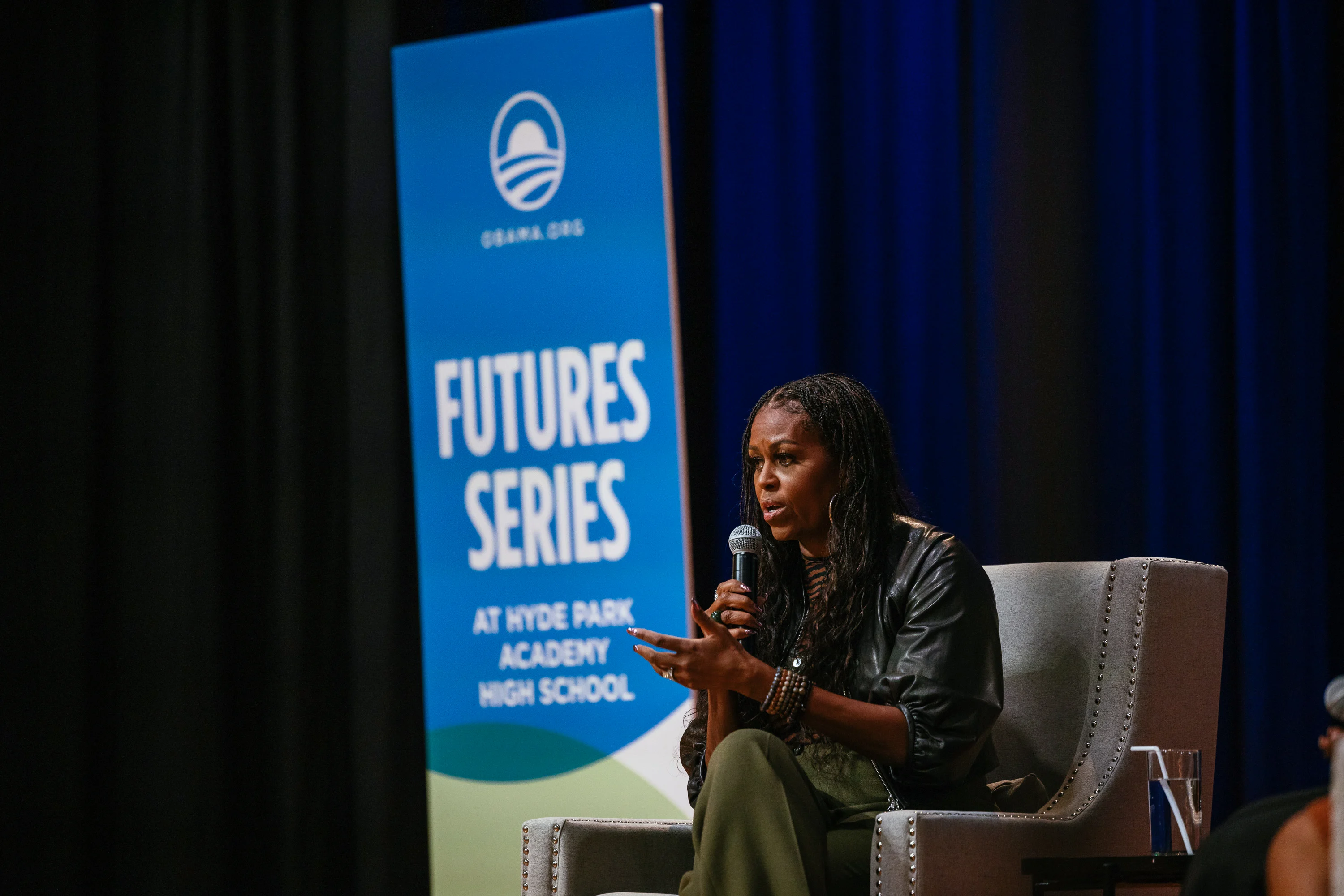 Former First Lady Michelle Obama is seated while holding a microphone and sporting long box braids. She is seated in front of signage that says “Futures Series” with the Obama Foundation logo. 