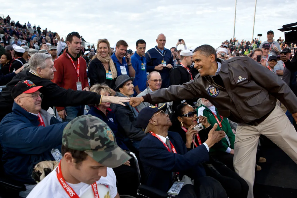 President Obama wearing a brown jacket and khaki pants leans over and fistbumps another man with a light skin tone wearing a black jacket. There is a crowd full of people with a variety of skin tones. They are all sitting outside. 