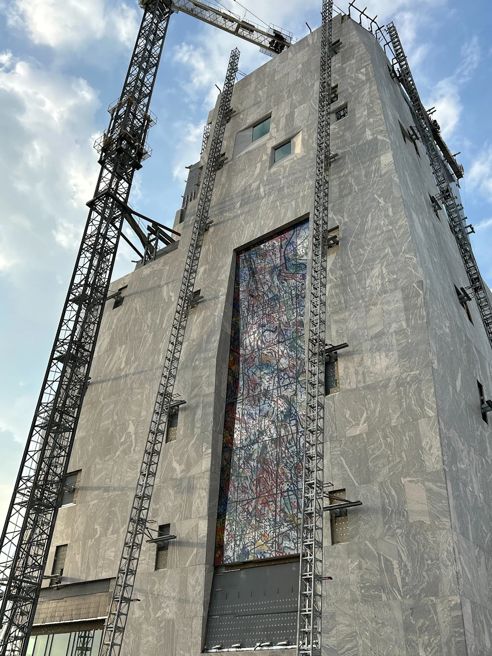 An upward image of the Obama Presidential Center Museum building during construction. The building is made of granite and stone. In the center of the building is "Uprising of the Sun," an art installation created by artist Julie Mehretu. Around the building are large scaffoldings. Behind the building are clouds.