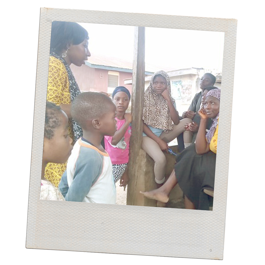 A Polaroid-style photo of a group of Black women and children dressed in various colors and patterned fabrics. Some sit on a concrete wall while others stand. Behind them are the roofs of two buildings.