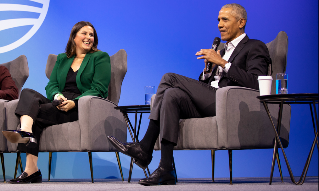 A photo of President Obama and a woman sitting in a grey comfy chairs on a stage. They sit infornt of a sky blue colored wall that has a portion of the Obama Foundation logo showing on the left. President Obama has a classic black suit, white button up, black socks, and black shoes on. The woman has light-medium skin tone, long brown hair, a green jacket, with a black jumpsuit underneath. She is holding a microphone down by her thigh while laughing with President Obama. Presidnet Obama is lightly laughing preparing to speak into his mic. 