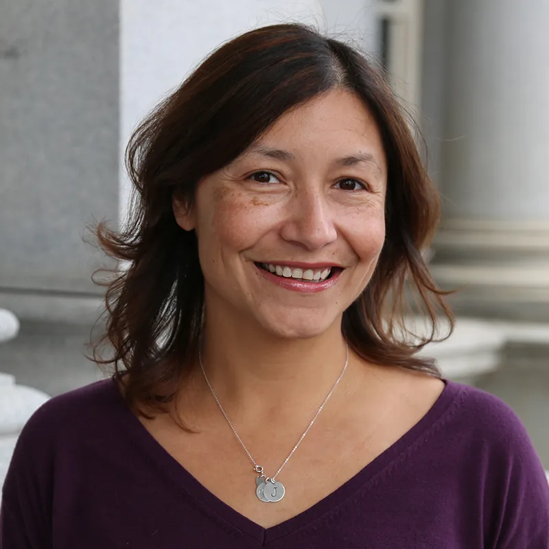 A headshot image of Julie Chávez Rodriguez. She has an olive complexion and dark hair that falls to her shoulders. She is smiling in the picture. She is wearing a purple shirt with a v-neck collar and a pendant necklace with three circles. One of the circles has the letter "J." She is standing outside.