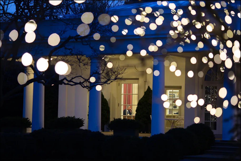 President Barack Obama is seen from the Rose Garden as he works at his desk  in the Oval Office following a meeting with the bipartisan, bicameral leadership of Congress for discussions on the fiscal cliff and a balanced approach to the debt limit and deficit reduction, in the Oval Office, Dec. 28, 2012. Lights illuminate the Rose Garden.  (Official White House Photo by Chuck Kennedy)