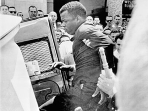 A black and white photo of a man with a deep skin tone is forced into a car as a crowd of other men with light skin tones watches 