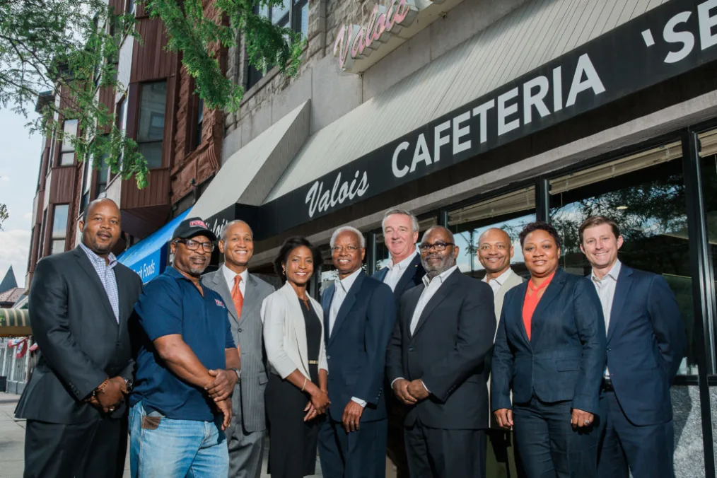 A group of men and women with different skin tones, most wearing suits, pose in front of a storefront with the words "Valois Cafeteria" on its sign.