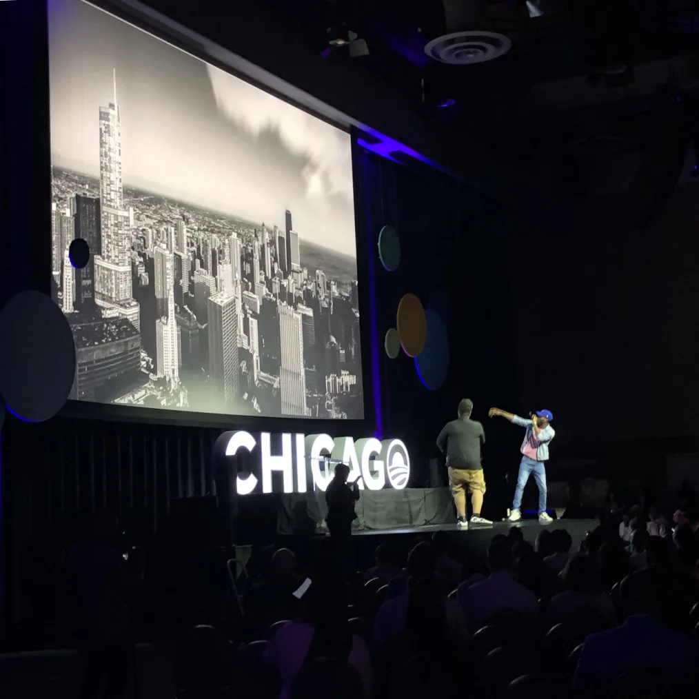 2 men stand on a stage with a medium to deep skin tone. One with a colorful attire and the other is black top and tan bottoms. Behind them is a large screen displaying a city with a sign that reads "Chicago" at the bottom 