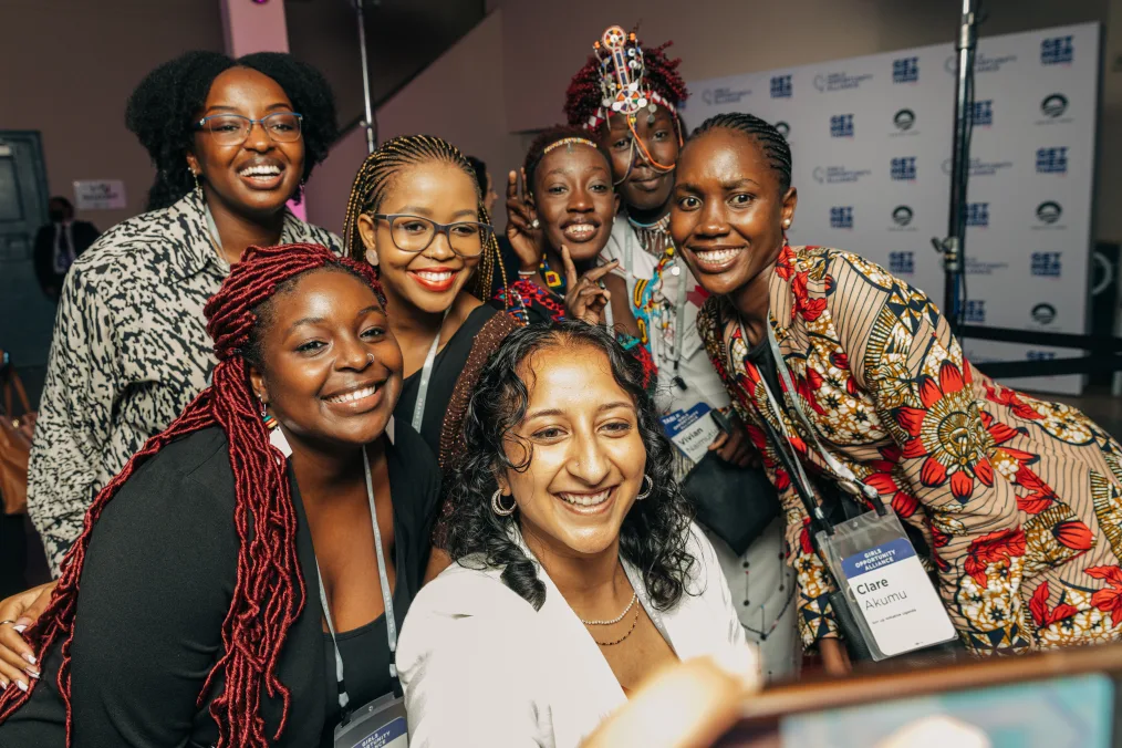 A selfie of a group of smiling middle aged woman with medium-deep to deep skin complexion. They are wearing formal attire and dresses. Some of them have cultural decorative pieces on. Most of them have lanyards with their names. The only clearly visible full name on the right is "Clare Akumu."  