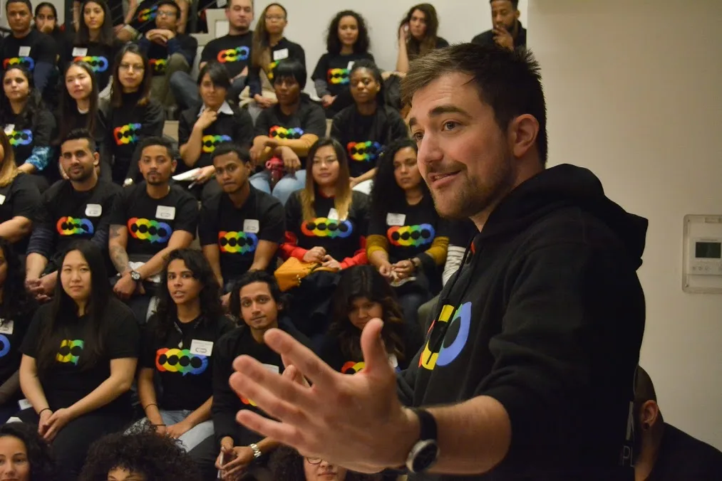 A man with spiky brown hair and beard stands in the front-right of the frame with his hands reaching out. Behind him are people all wearing the same t-shirt featuring red, yellow, green and blue circles and sitting in rows.
