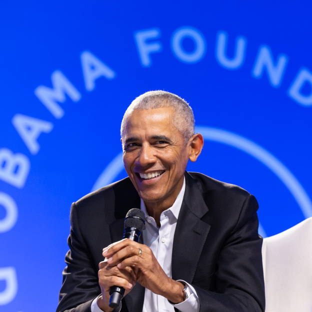 President Obama holds a microphone as he sits on a stage at the 2024 Democracy Forum. He is smiling. A blue sign in the background reads, “Obama Foundation.” 