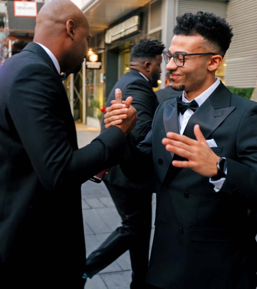 A Black man with a light skin tone shakes hands with a Black man with a medium skin tone. Both are tuxedos.