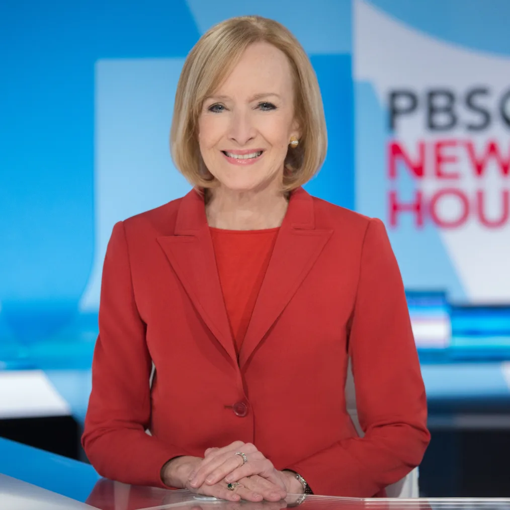 A White woman with blonde, chin-length hair wearing a red suit smiles with her teeth showing. Her hands on rest, one on top of the other, on a desk. Behind her is a blue geometric pattern and part of the worlds "PBS News Hour." 