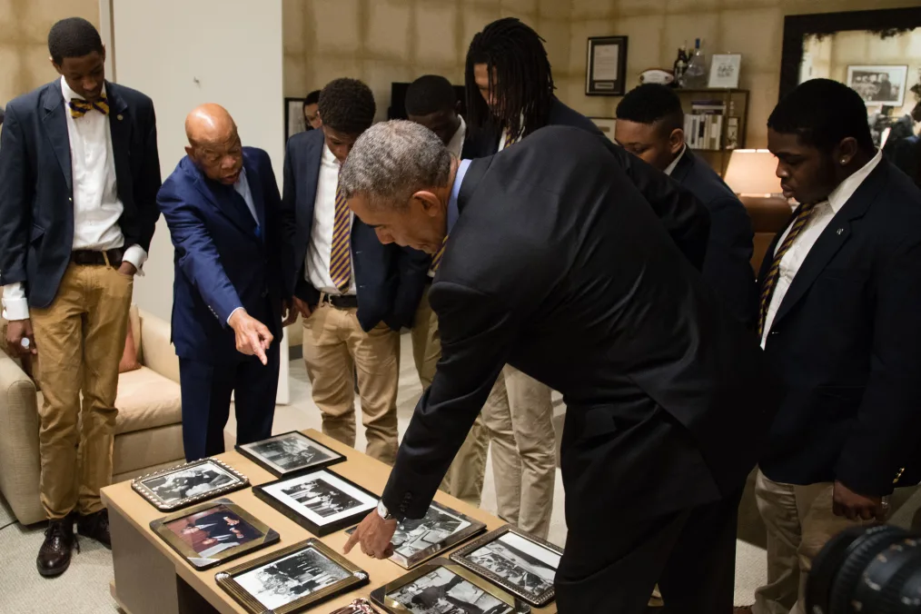 Barack Obama, dressed in a dark suit, points with his left hand to one of many photos sitting on a light colored coffee table. Across from him John Lewis, dressed in a blue suit, is using his right hand to point to the same photo. Six young Black men dressed in dark jackets, white shirts and brown pants surround the two men looking at the photos.