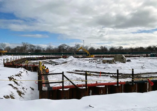 Snow covers a construction site. A yellow digger can be seen in the background.