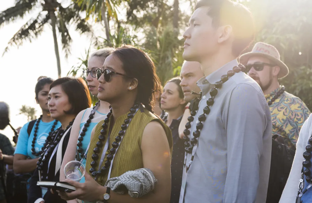 A group of people wearing large-bead necklaces look to the left. Palm trees are behind them.