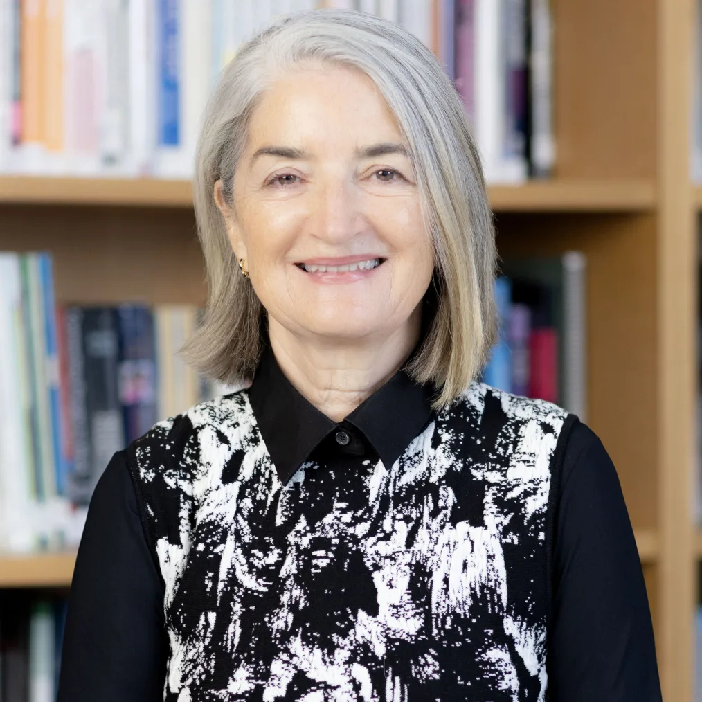 A portrait of a light-skinned woman with gray hair. She smiles showing her teeth and wears an abstract print black and white collared shirt. Behind her are books on a book shelf.