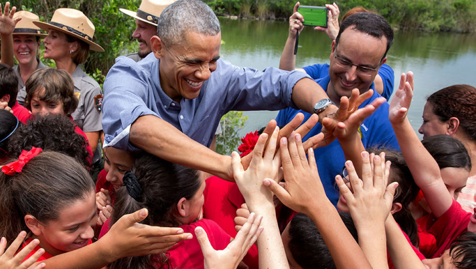 Barack Obama reaches out his hands to high-five a group of children wearing red t-shirts. Behind him is a body of water. 