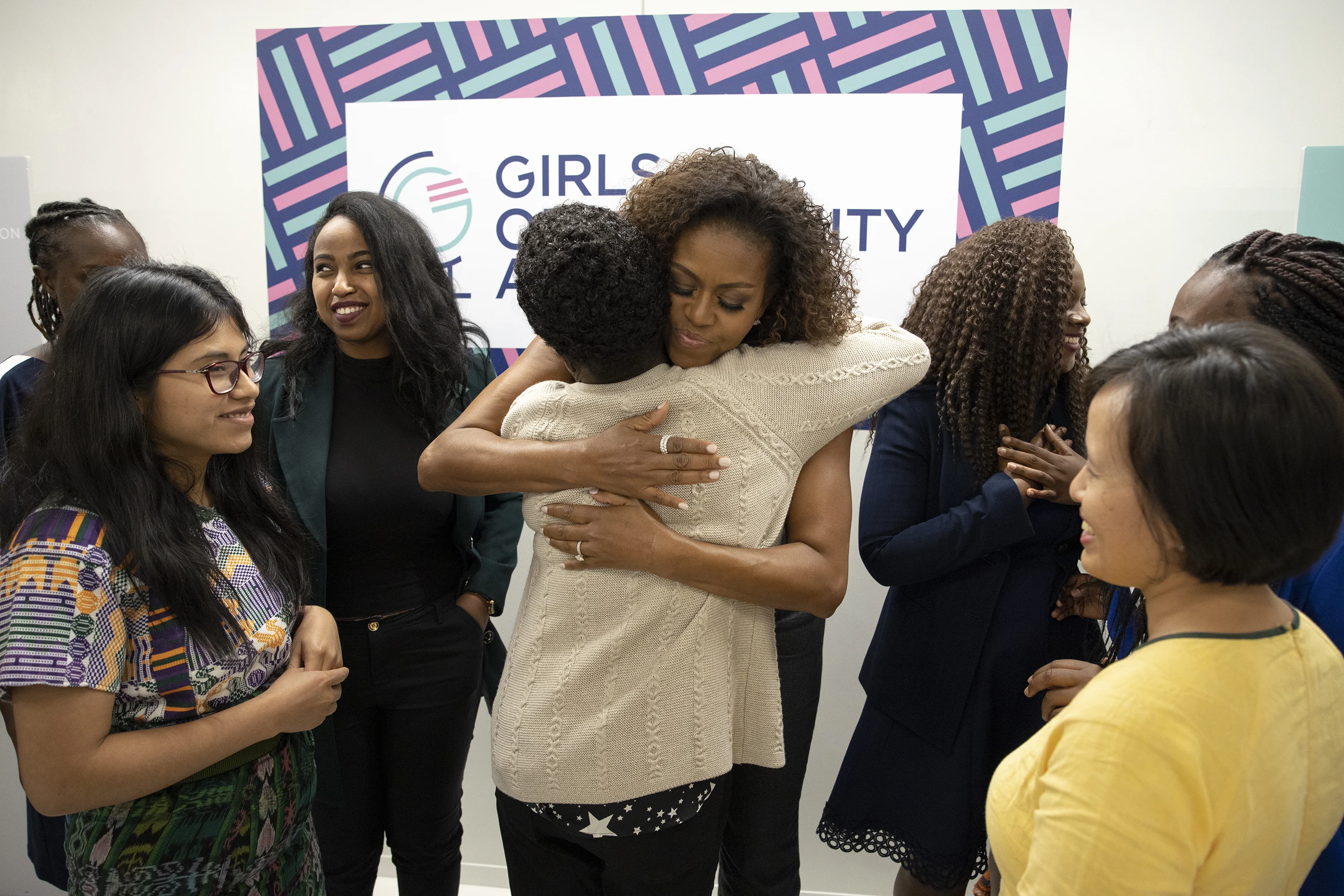 There are three women on the right side of the image and three women on the left side of the image. In the center of the image, Michelle Obama is hugging a woman and facing the camera. Michelle Obama has curly hair and is wearing several rings. The woman she is hugging is facing opposite the camera – we cannot see her face – she has short curly hair and is wearing a beige sweater and black pants. In the background, several women with a range of light to deep skin tones and ages speak to one another. On the wall of the room is a poster that reads, “Girls Opportunity Alliance.” The poster has a border with short perpendicular lines in pink and light blue with a dark blue background. 