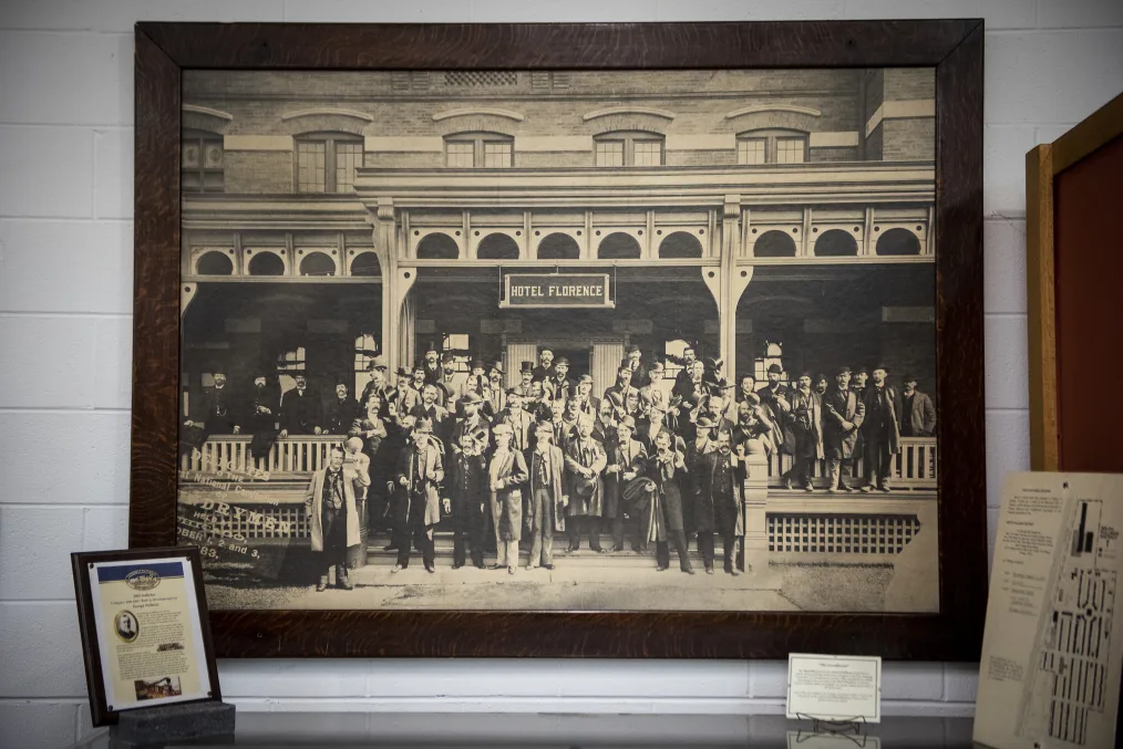 A picture of a framed picture of a large group of white men with large mustaches and beards, suits, and bowling hats. They stand at the stairs and landing of a building which hanging sign above reads "Hotel Florance". 