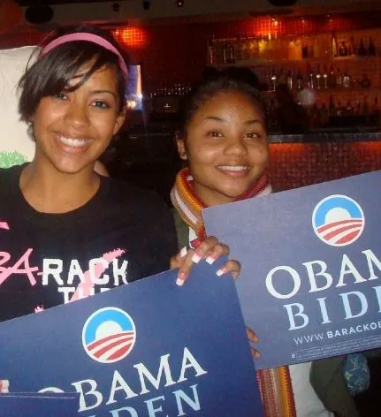 Two smiling medium-dark skin toned women hold up "Obama Biden" signs. 