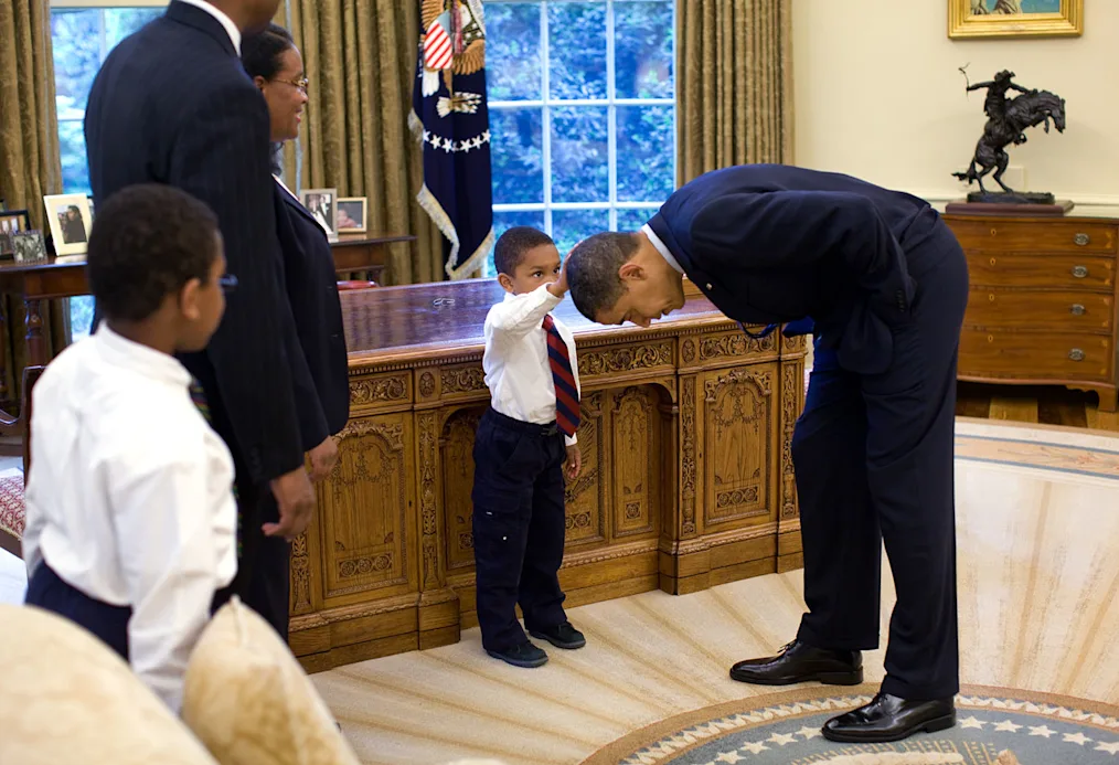 A young Black boy reaches out to touch President Obama's hair. They are in the oval office and a two other adults and one child stand to the side.