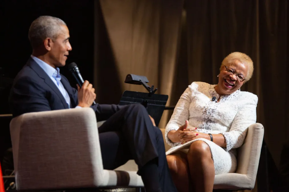 President Obama sits on a stage in a chair with a woman with medium deep skin tone, short blond hair and glasses   