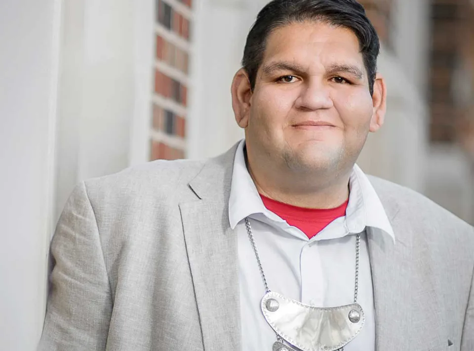 Corey Still, a man with a light-medium skin tone and combed over black hair wearing a light grey suit jacked and white collared shirt, looks into the camera.