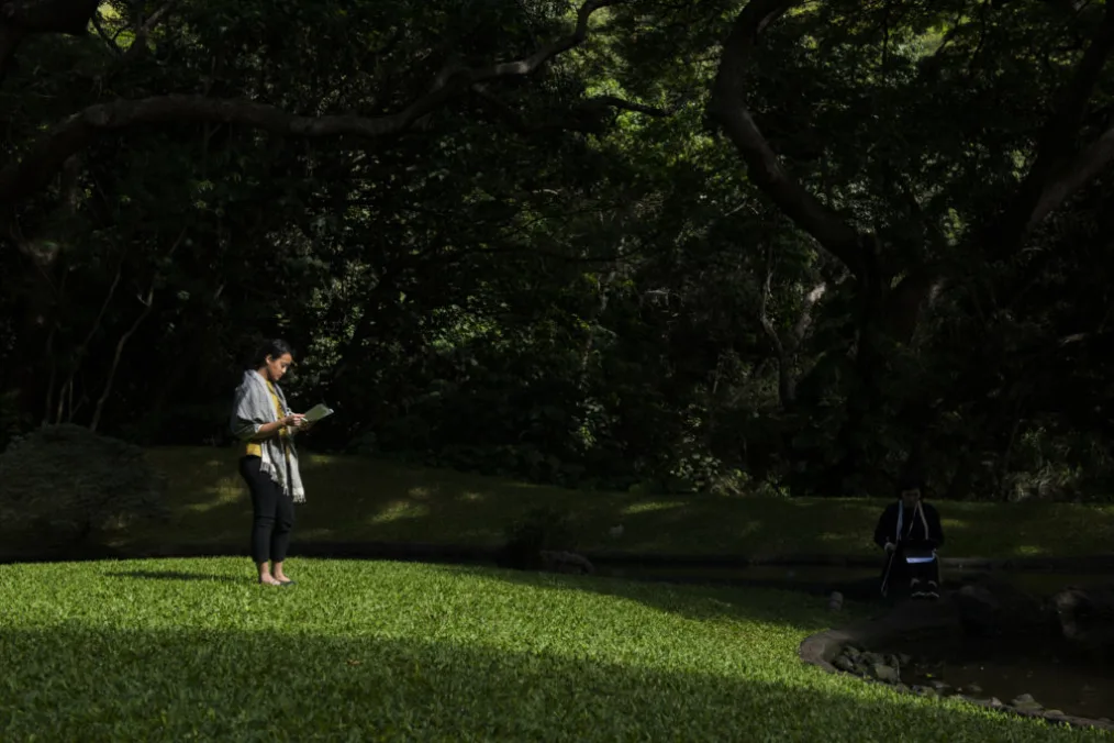 From a distance, a woman with a medium skin tone stands in a dimmed forest-like atmosphere, reading a book.