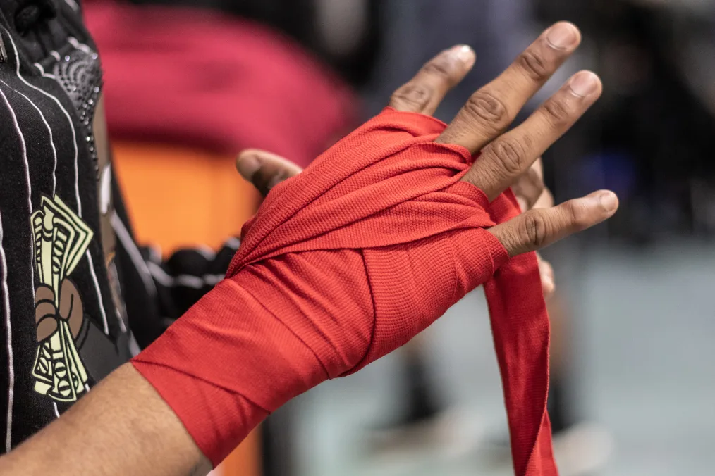 A arm and hand, that is medium-deep toned, in focus of the camera. The hand is being wrapped in red boxing tape.