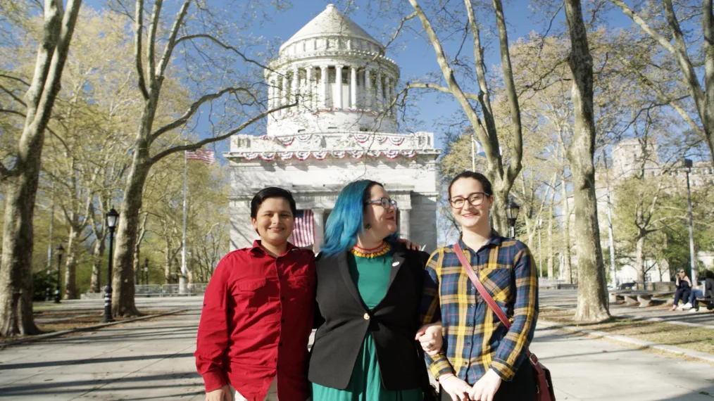 A young boy with a light skin tone and black hair wears a red shirt. Next to him is a woman with a light skin tone, blue hair, and glasses wearing business attire. Lastly, another woman with a light skin tone and red glasses wears a plaid shirt. The three of them stand in front of a historic building smiling. 