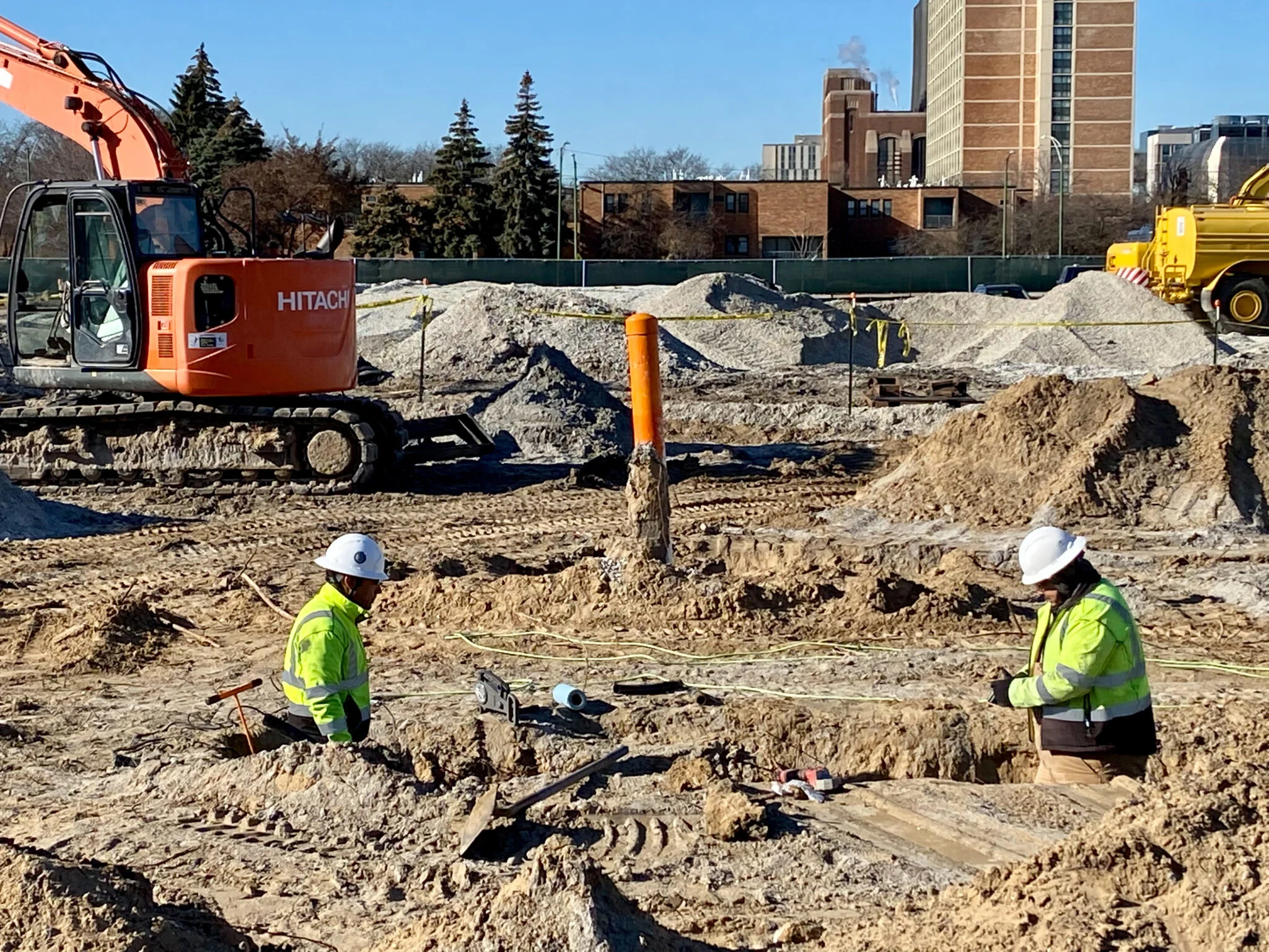 Two men in neon yellow construction jackets and hardhats stand waist-deep in a dirt trench. Behind them are construction vehicles and a blue sky with buildings.