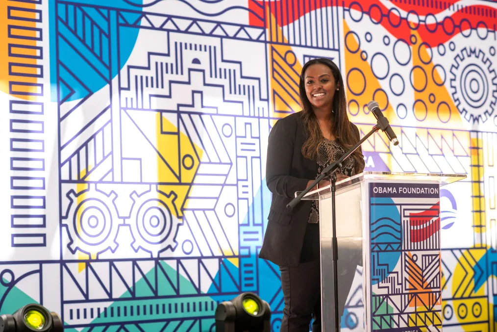 A young woman smiles at the crowd while speaking at a podium in front of a multicolored, graphical background pattern.