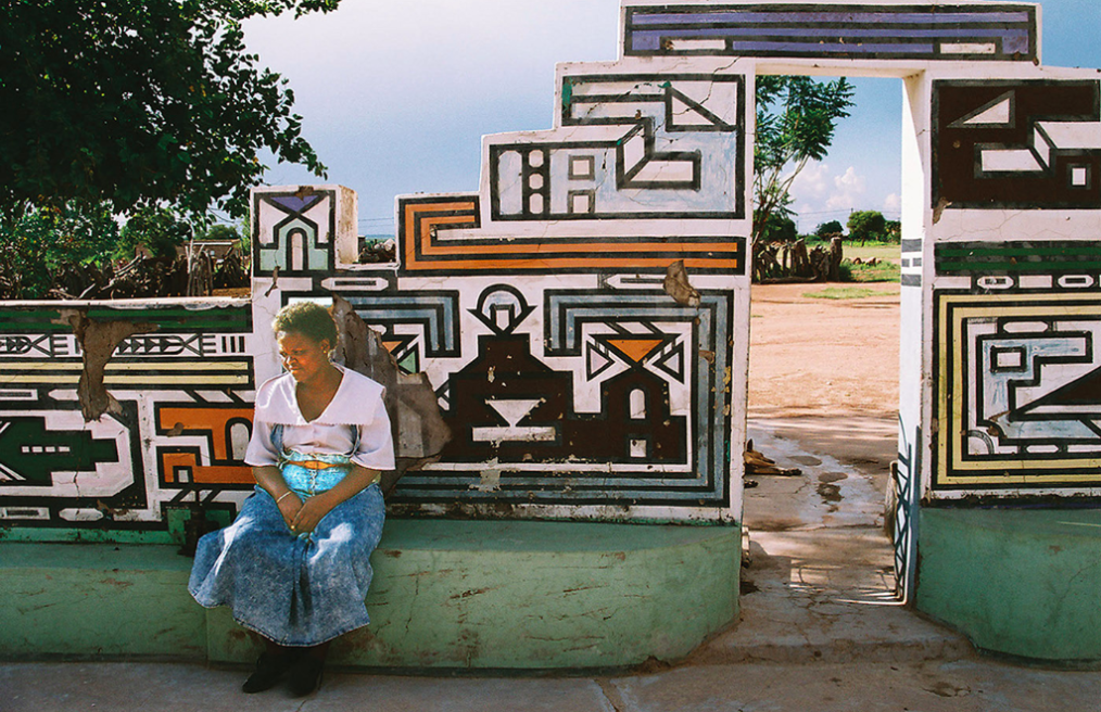 A woman sits in front of a wall structure with geometric shapes and designs.