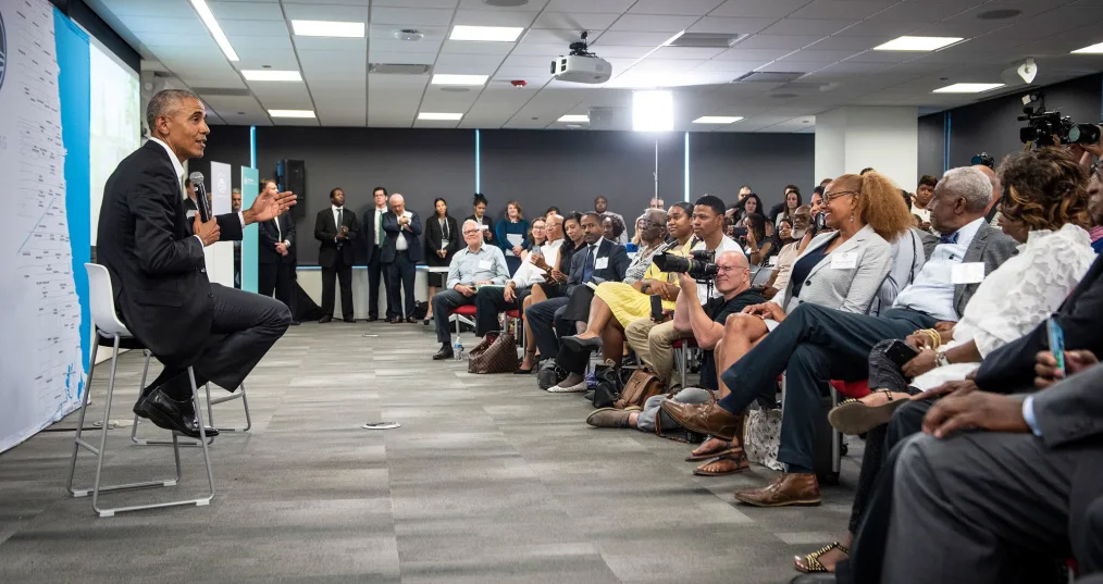 President Obama giving a presentation to a large group of people of medium to deep skin tones. President Obama is seated in a small chair. To his left is a projecting screen, a map, and Obama Foundation signs.